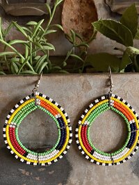 Colorful beaded earrings hang against a rustic metal background. The beads form a loop with alternating sections of green, yellow, red, blue, white, and black. The earrings have a vibrant and artistic design.