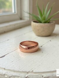 A shiny copper ring rests on a white, rustic wooden table near a small potted succulent and a sunlit window. The KVK BOHOFAIR logo appears in the bottom right corner.