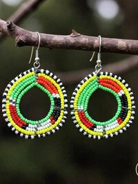 A pair of colorful beaded hoop earrings in red, yellow, green, black, and white hang from a tree branch. The earrings have intricate circular patterns and are photographed outdoors.