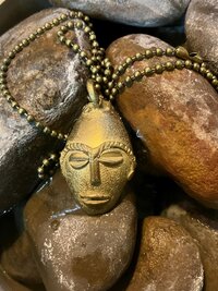 A brass pendant necklace with a stylized face design rests on smooth, wet stones. The pendant is attached to a beaded chain
