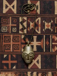 A beaded necklace with a metallic, stylized African mask pendant is laid on a woven fabric featuring geometric tribal patterns in earthy tones. The pendant is centered on the patterned textile background.