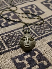 A metallic pendant shaped like an abstract face rests on a patterned fabric with geometric designs. The pendant hangs from a beaded chain.