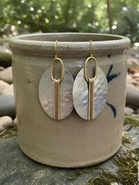 A pair of silver oval earrings with a hammered texture, accented with gold-colored metal and a vertical copper-colored stripe. The earrings hang from gold-colored ear wires. The earrings are displayed in front of a ceramic pot.