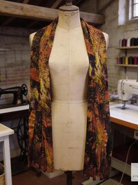 A colorful tie dye scarf with in shades of  brown, orange, and green is draped on a mannequin. The scarf is the focal point, showcasing a unique design. The background includes sewing equipment.