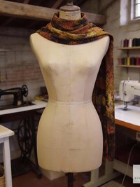 A patterned tie dye scarf in shades of brown, orange, and green is draped over a dress form. The scarf's design features a mix of floral and abstract elements. The dress form is in a workshop setting, with sewing machines and spools of thread visible in the background.