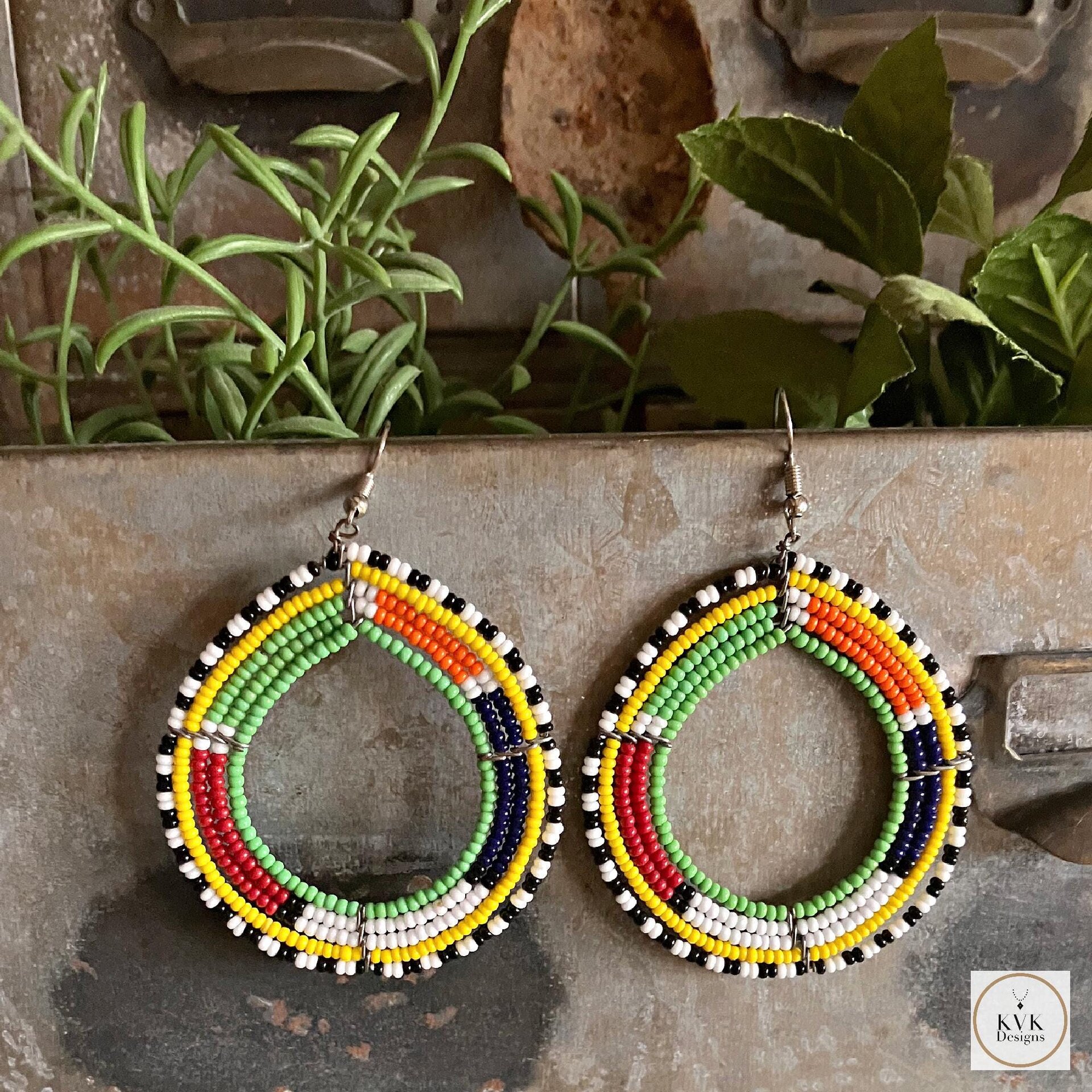 Colorful beaded earrings hang against a rustic metal background. The beads form a loop with alternating sections of green, yellow, red, blue, white, and black. The earrings have a vibrant and artistic design.