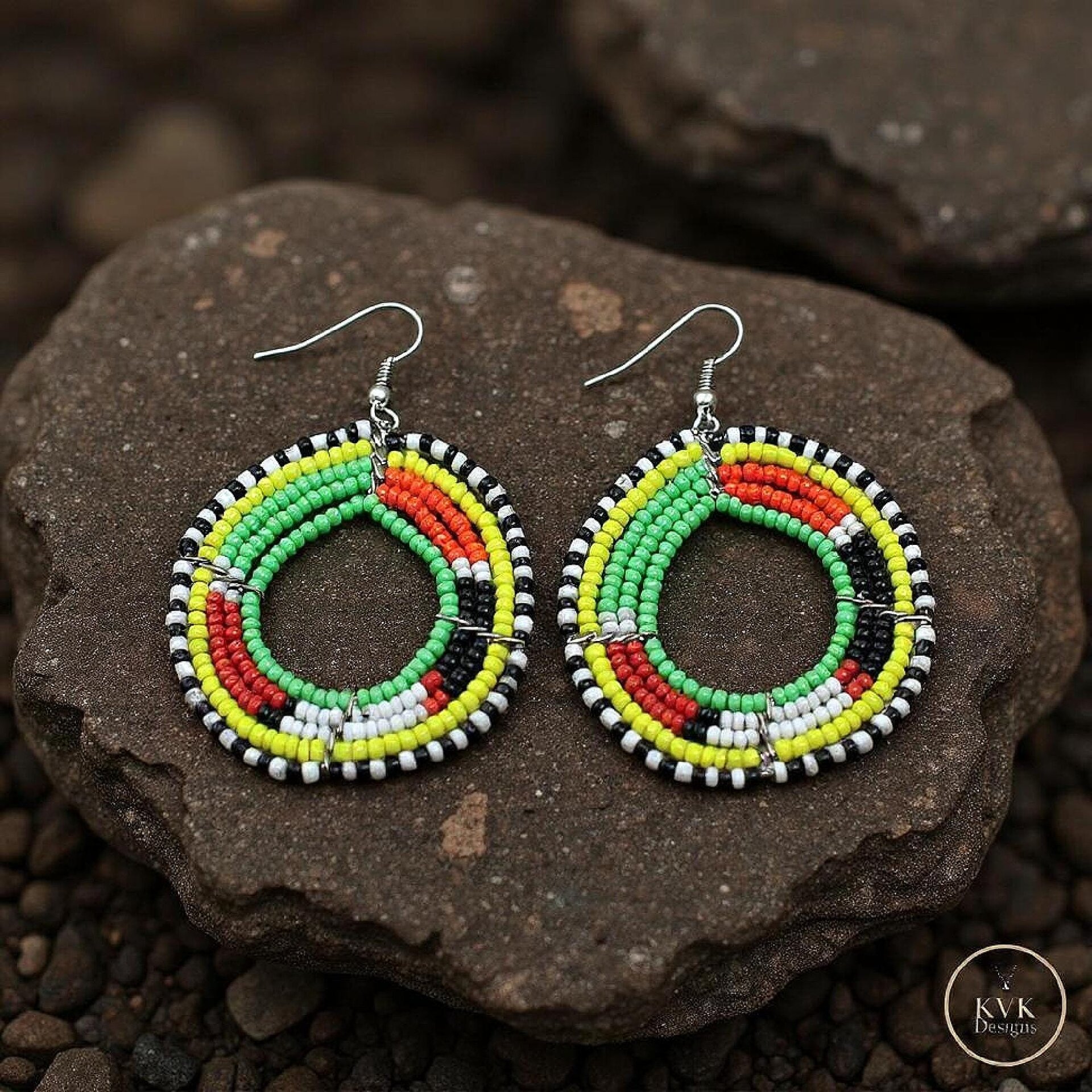 A pair of colorful beaded hoop earrings with green, yellow, red, white, and black beads arranged in concentric patterns, displayed on dark brown textured stones.