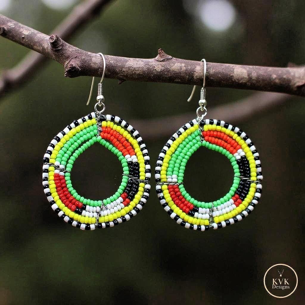 A pair of colorful beaded hoop earrings in red, yellow, green, black, and white hang from a tree branch. The earrings have intricate circular patterns and are photographed outdoors.