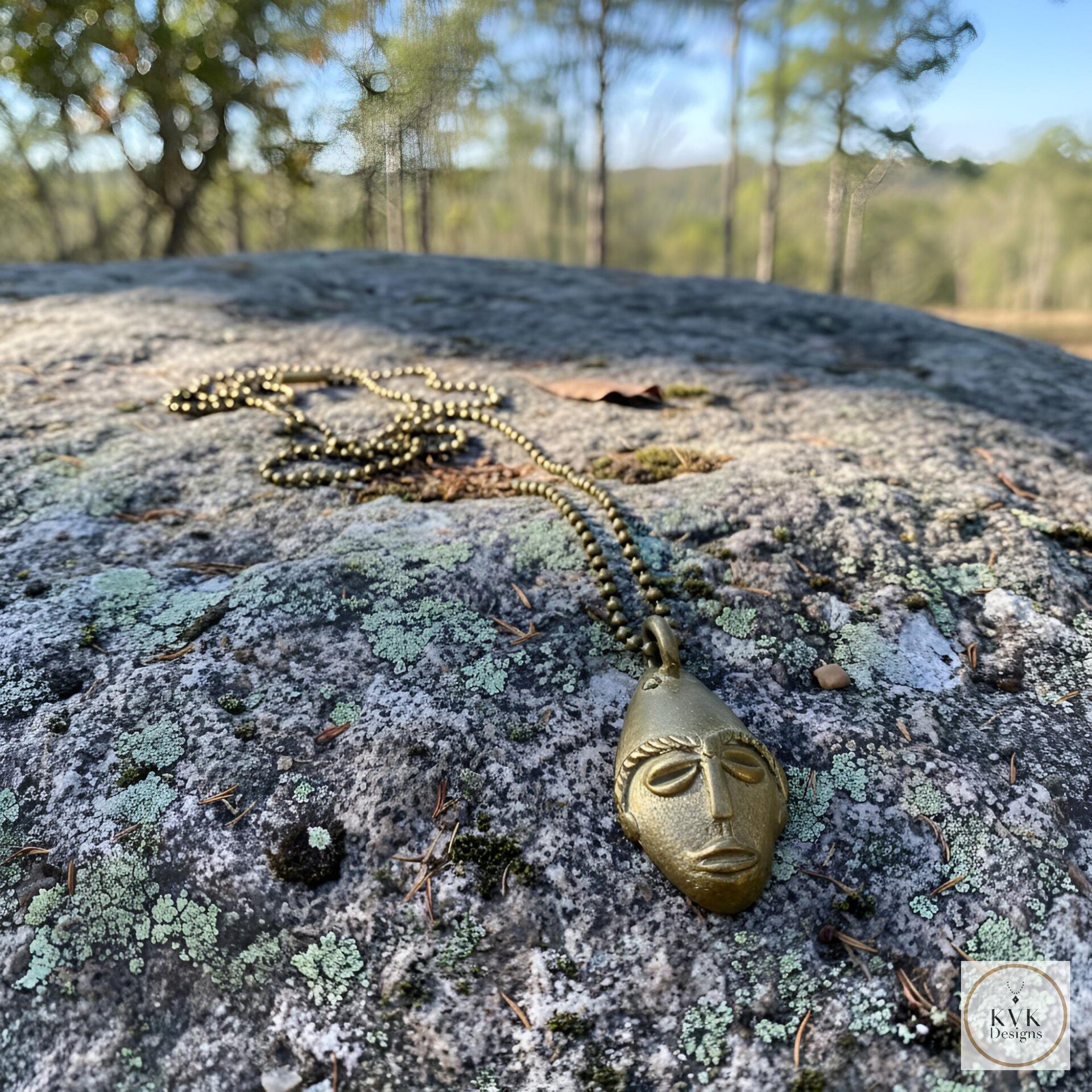 A brass pendant necklace with a sculpted face rests on a lichen-covered rock outdoors, with blurred green trees and hills in the background.
