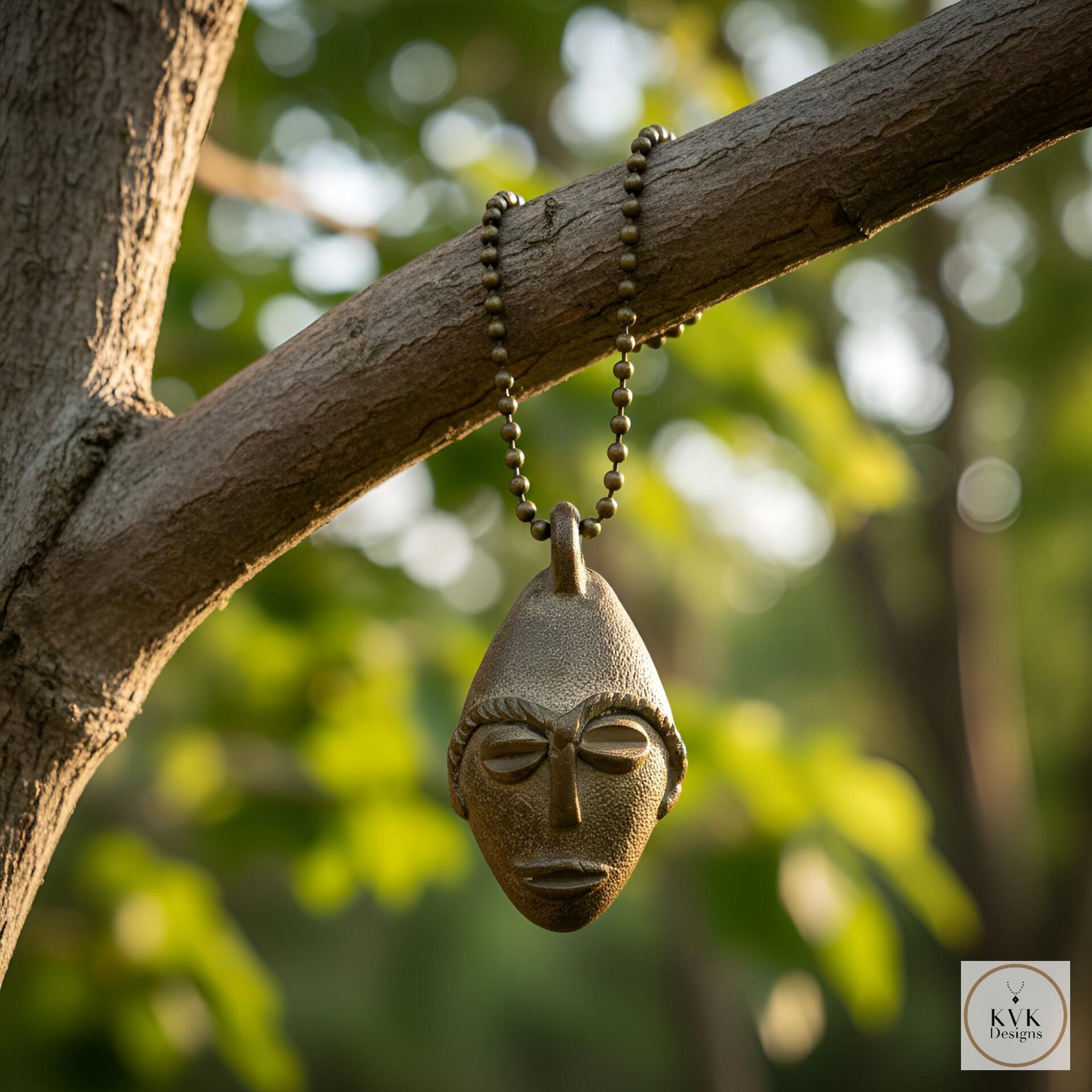 A brass pendant shaped like a stylized human face hangs from a chain on a tree branch, with sunlight filtering through green leaves in the blurred background.