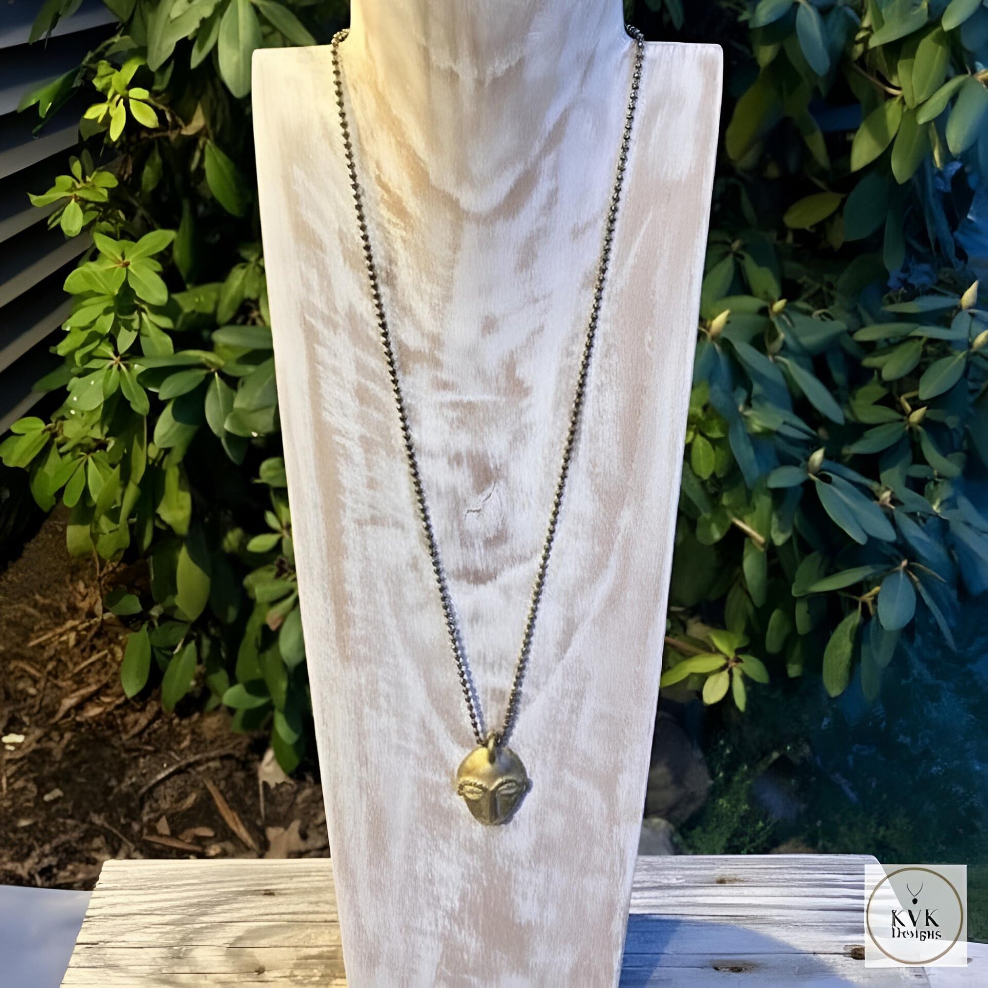 A metallic necklace with a sculpted face pendant is displayed on a light wooden bust, set outdoors with green foliage in the background.