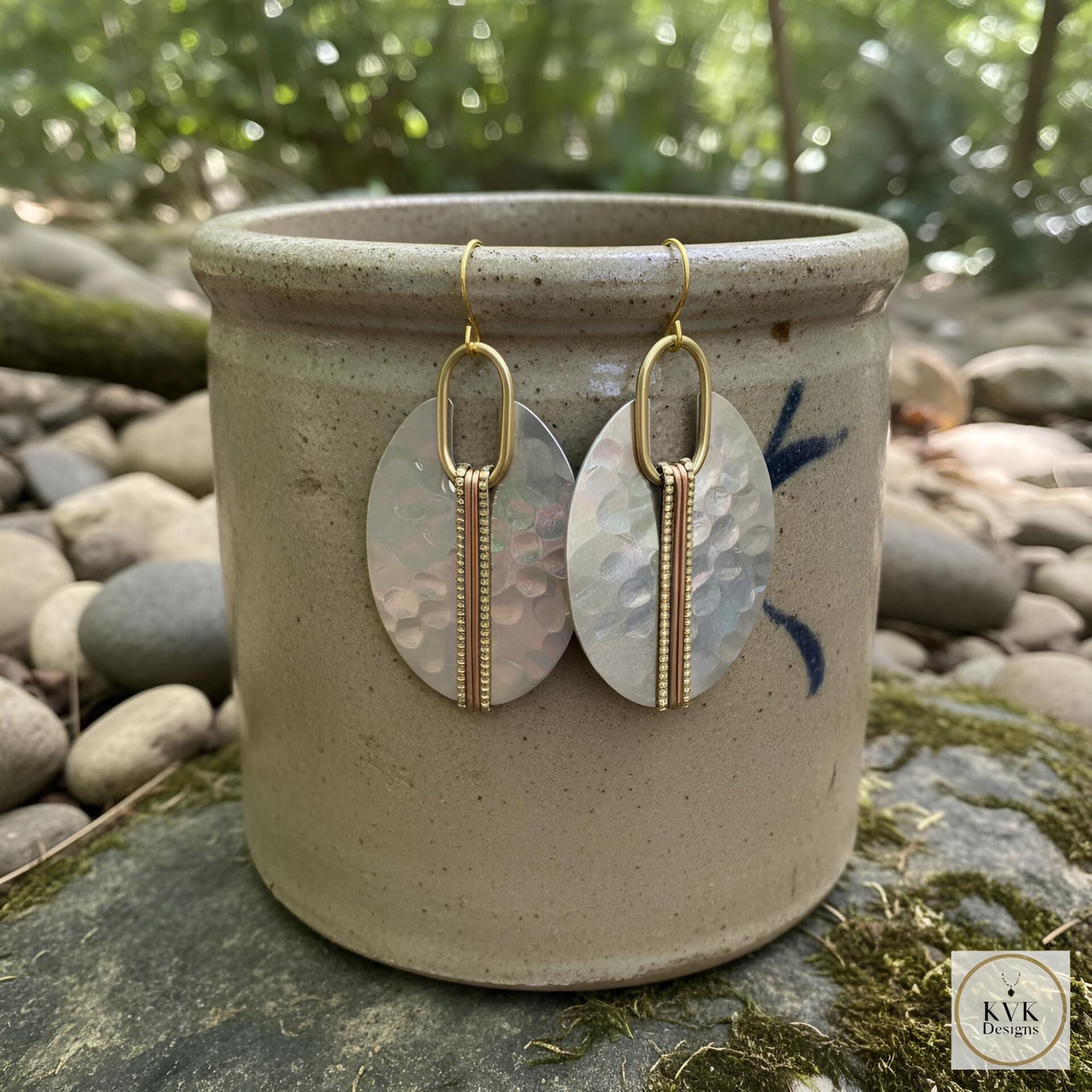 A pair of silver oval earrings with a hammered texture, accented with gold-colored metal and a vertical copper-colored stripe. The earrings hang from gold-colored ear wires. The earrings are displayed in front of a ceramic pot.