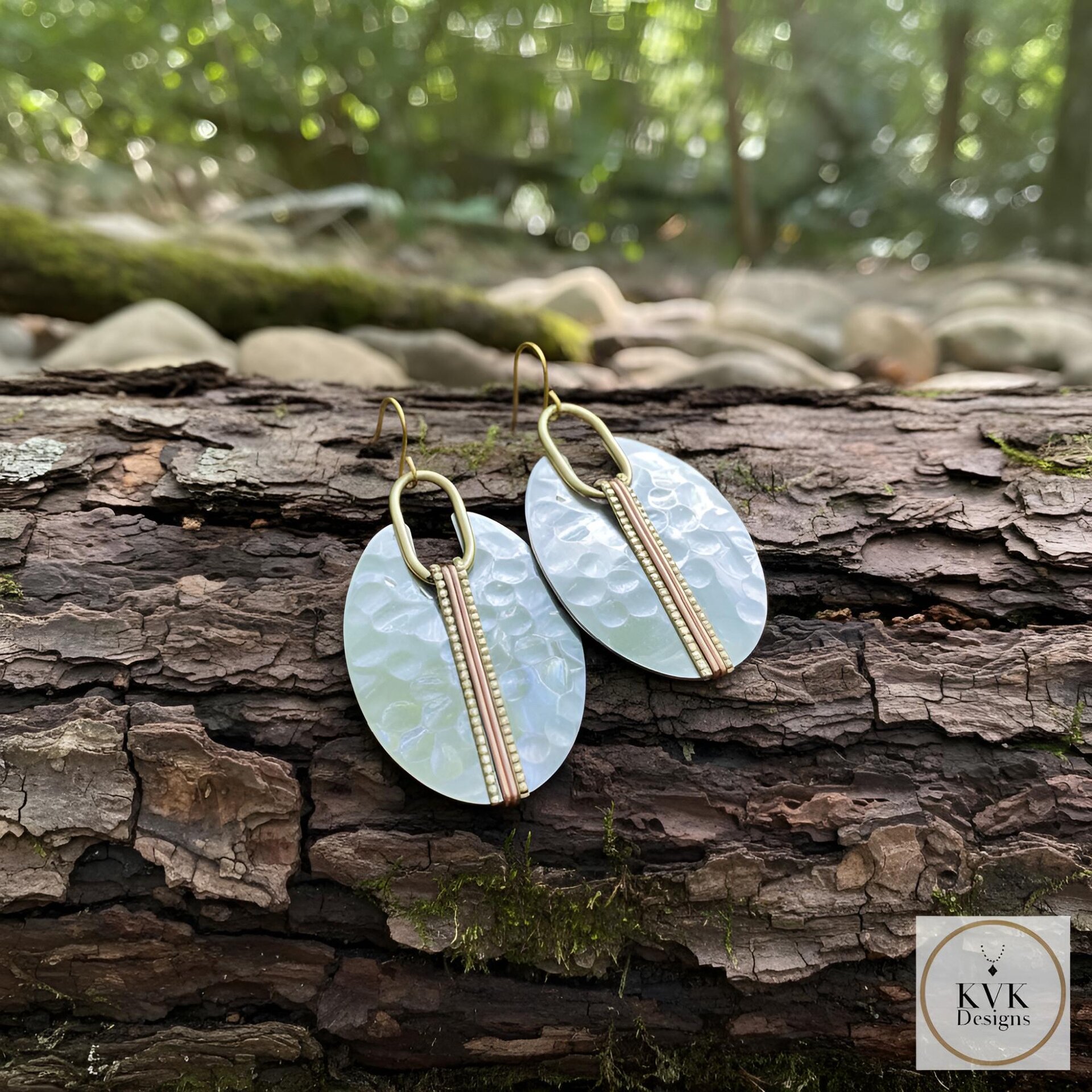 A pair of oval-shaped earrings with a hammered silver finish. Each earring features a vertical copper-colored stripe down the center, accented with small gold beads. The earrings hang from gold-colored ear wires. The background is a natural setting.