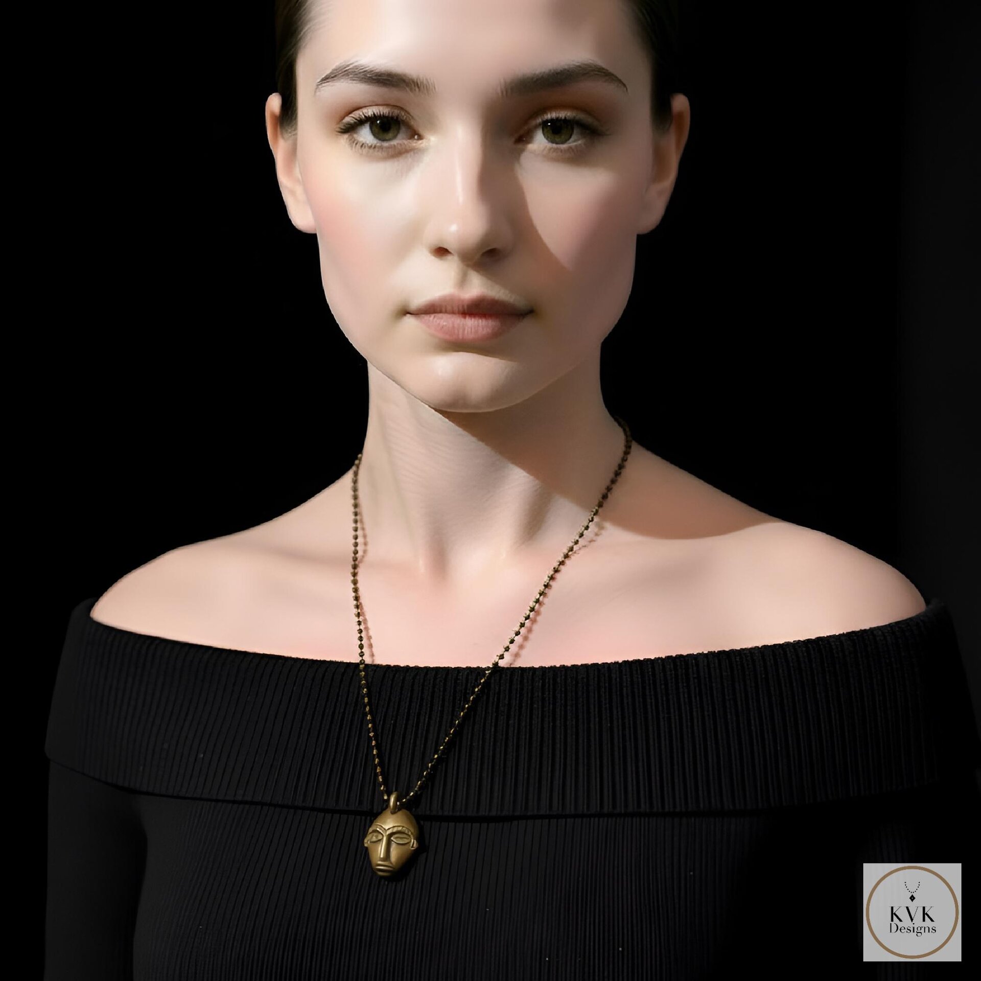 A woman in an off-the-shoulder black top wears a gold pendant necklace shaped like a face. She has light skin, tied-back dark hair, and poses against a black background.