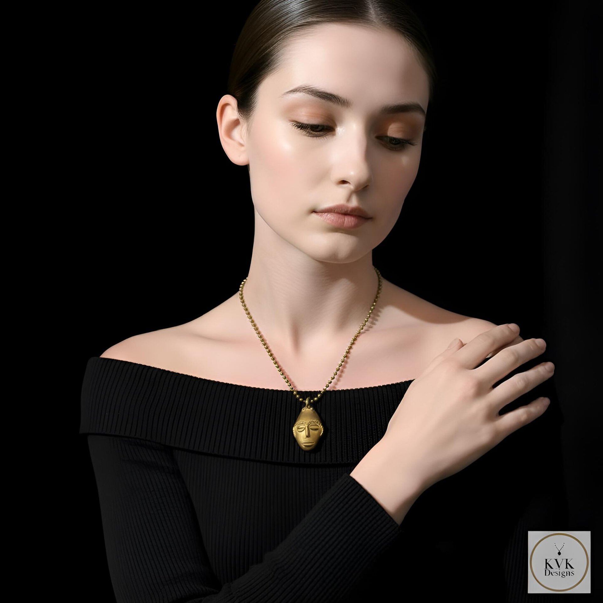 A woman wearing an off-shoulder black top and a gold pendant necklace with a sculpted face design poses against a black background, gently touching her shoulder.