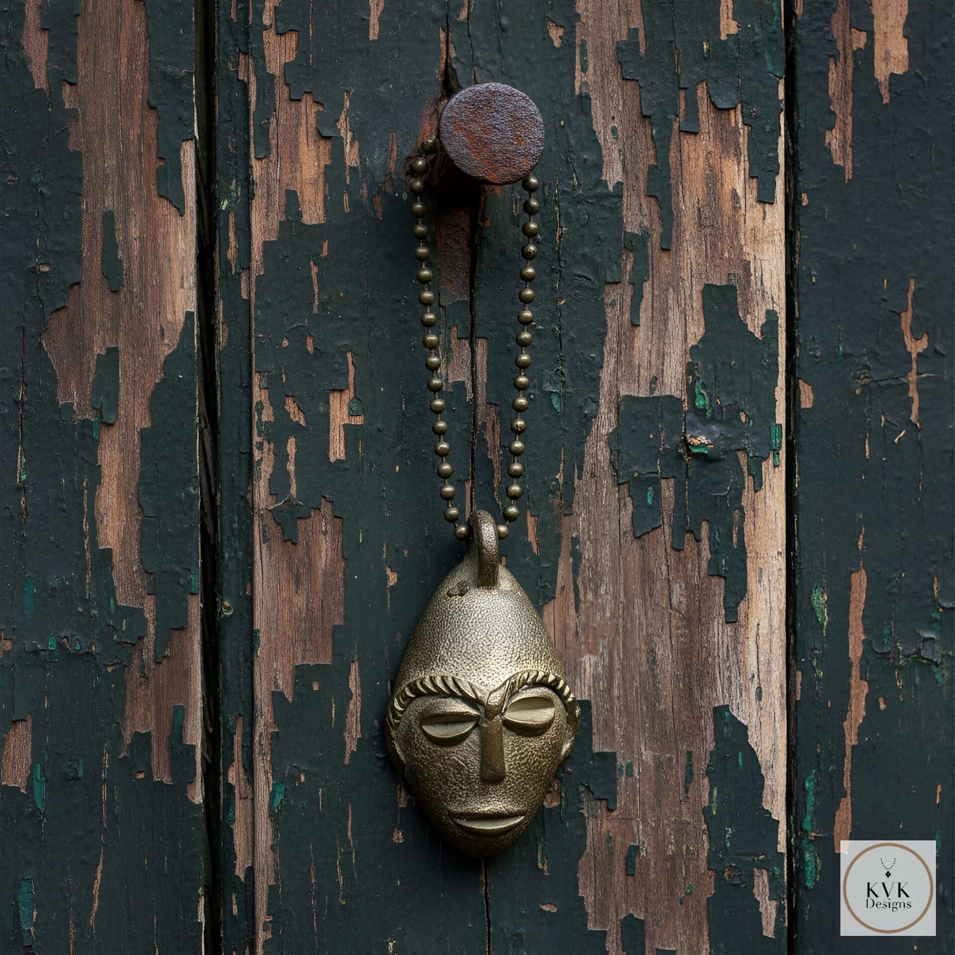 A bronze pendant shaped like a stylized human face hangs from a metal ball chain on a weathered, peeling wooden surface.