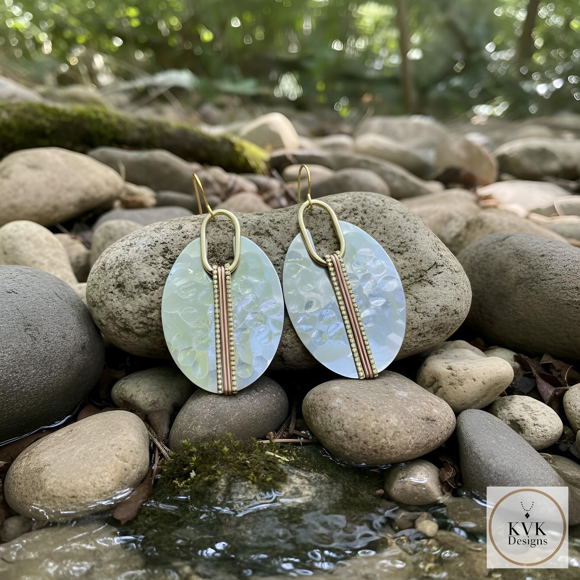 Oval-shaped earrings with a pearlescent finish, featuring a vertical copper and gold accent. The earrings have gold-colored ear wires and are displayed on a rock. The earrings are a fashion accessory.