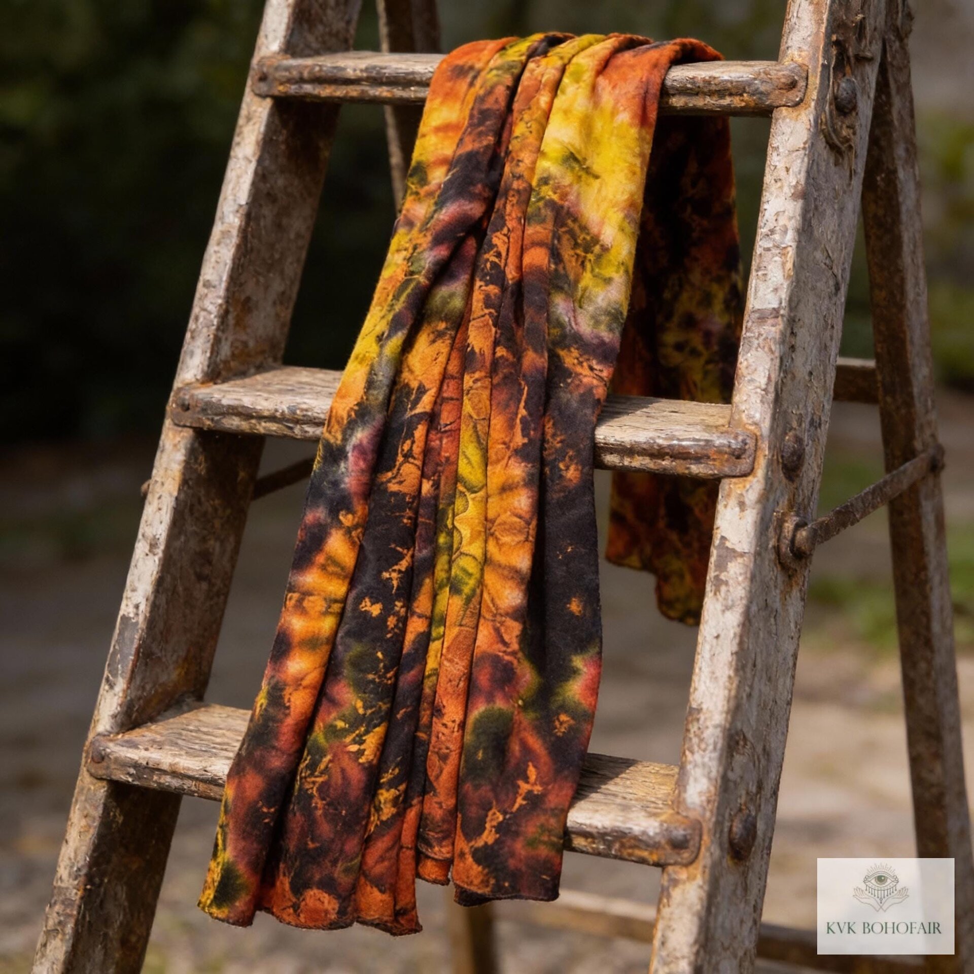 A tie-dye scarf in shades of orange, yellow, and black is draped over a rustic wooden ladder. The scarf's pattern features a blend of colors, creating a vibrant and artistic design. The ladder's aged appearance adds a vintage touch to the scene.