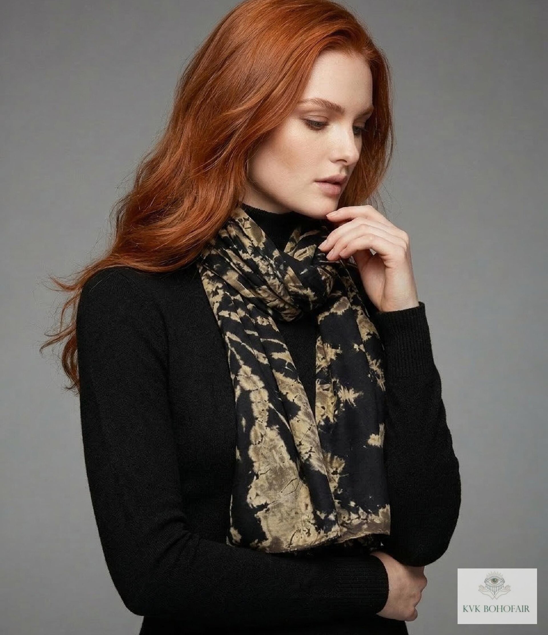 A woman with long red hair wears a black turtleneck sweater and a black and beige patterned scarf. She stands against a neutral gray background, looking down with one hand near her chin. Logo "KVK BohoFair" is in the lower right corner.