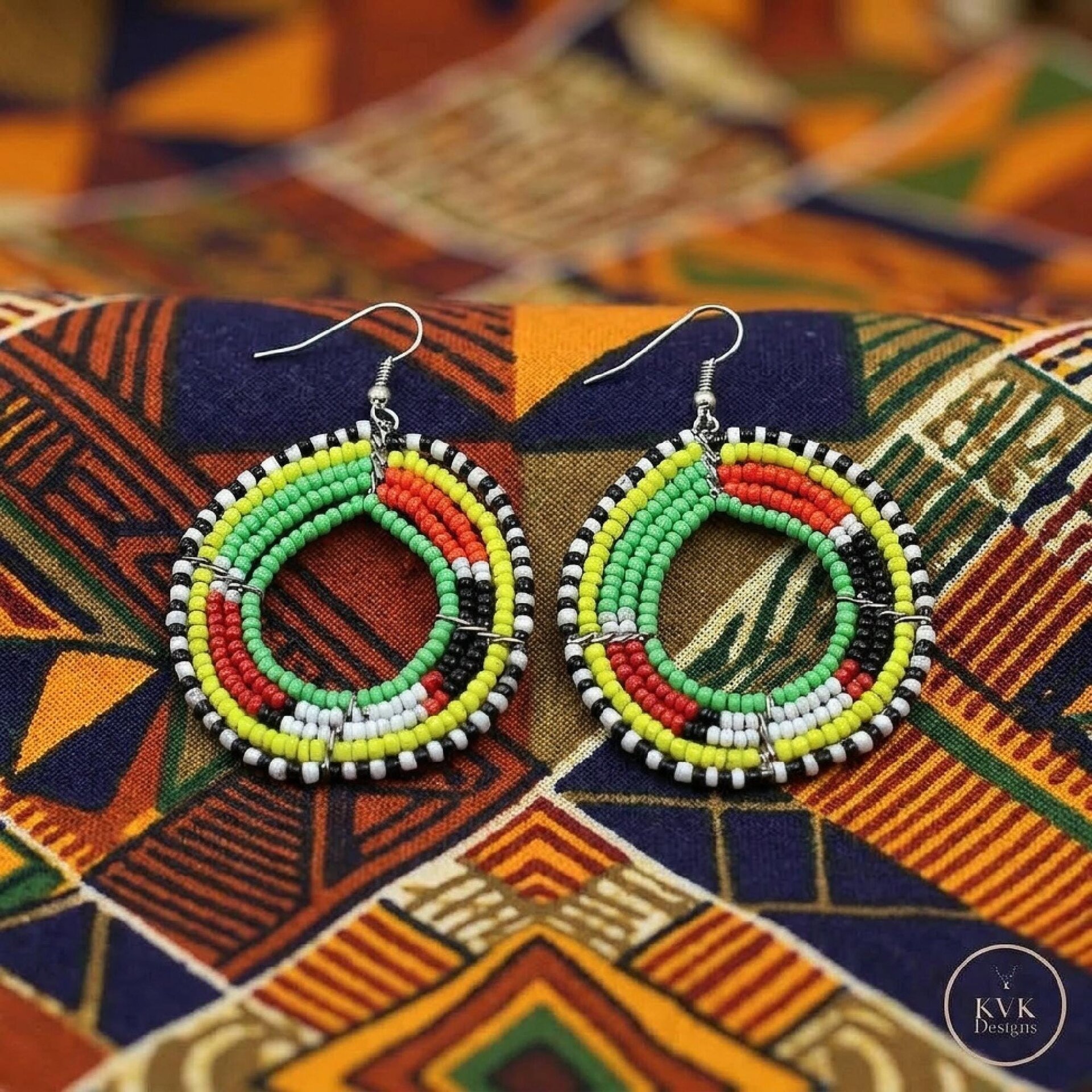 A pair of colorful beaded hoop earrings with green, yellow, white, red, and black beads, displayed on vibrant geometric-patterned fabric.