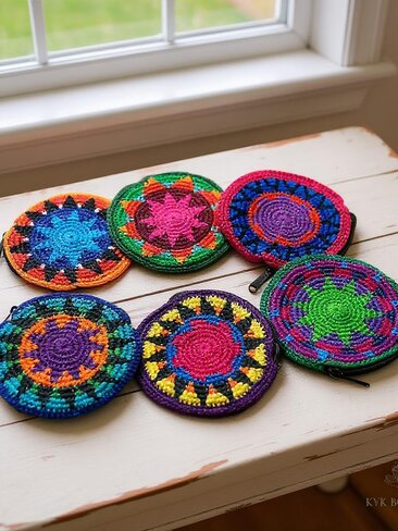 Six colorful, round, crocheted coin purses with intricate geometric patterns are displayed on a white wooden table near a window. Each coin purse features different vibrant color combinations and designs.