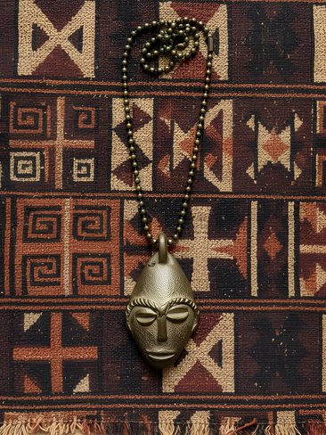 A beaded necklace with a metallic, stylized African mask pendant is laid on a woven fabric featuring geometric tribal patterns in earthy tones. The pendant is centered on the patterned textile background.