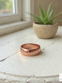 A shiny copper ring rests on a white, rustic wooden table near a small potted succulent and a sunlit window. The KVK BOHOFAIR logo appears in the bottom right corner.