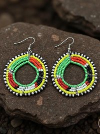 A pair of colorful beaded hoop earrings with green, yellow, red, white, and black beads arranged in concentric patterns, displayed on dark brown textured stones.