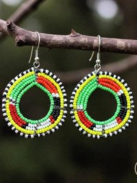 A pair of colorful beaded hoop earrings in red, yellow, green, black, and white hang from a tree branch. The earrings have intricate circular patterns and are photographed outdoors.