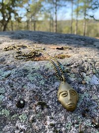 A brass pendant necklace with a sculpted face rests on a lichen-covered rock outdoors, with blurred green trees and hills in the background.