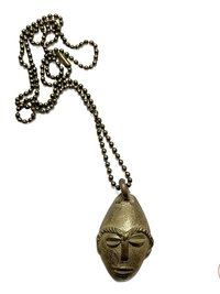 A brass necklace with a ball chain and a pendant shaped like a stylized, elongated face with distinct facial features, displayed on a white background.