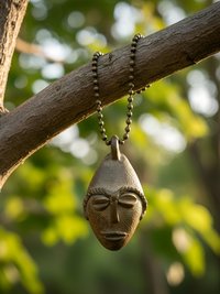 A brass pendant shaped like a stylized human face hangs from a chain on a tree branch, with sunlight filtering through green leaves in the blurred background.