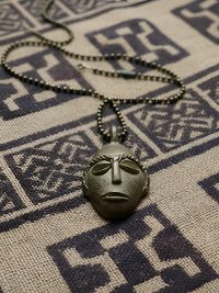 A metallic pendant shaped like an abstract face rests on a patterned fabric with geometric designs. The pendant hangs from a beaded chain.