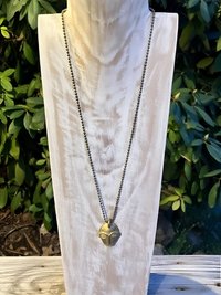 A metallic necklace with a sculpted face pendant is displayed on a light wooden bust, set outdoors with green foliage in the background.