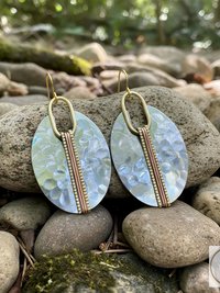 Oval-shaped earrings with a hammered silver finish. Each earring features a vertical copper-colored band down the center and gold-colored accents. The earrings are displayed on a bed of rocks, with a blurred natural background.