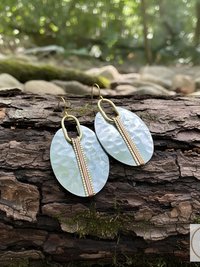 A pair of oval-shaped earrings with a hammered silver finish. Each earring features a vertical copper-colored stripe down the center, accented with small gold beads. The earrings hang from gold-colored ear wires. The background is a natural setting.