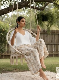 A woman sits in a macrame swing chair outdoors. She wears a white V-neck top and floral print pants. The swing hangs from a tree branch, and a rug is beneath the swing. The KVK Designs logo is in the bottom right corner.