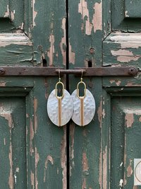 Oval-shaped earrings with a hammered texture and a gold-colored vertical accent. The earrings are suspended from gold-colored hooks. The background is a weathered green wooden door.