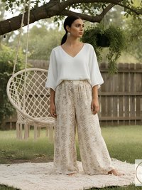 A woman wearing a white V-neck top and wide-leg pants with a gold floral pattern. The pants are cream-colored. The woman is standing on a cream-colored rug in a garden setting. A macrame swing chair hangs in the background.