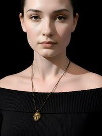 A woman in an off-the-shoulder black top wears a gold pendant necklace shaped like a face. She has light skin, tied-back dark hair, and poses against a black background.