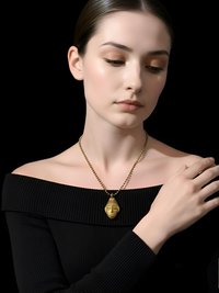 A woman wearing an off-shoulder black top and a gold pendant necklace with a sculpted face design poses against a black background, gently touching her shoulder.