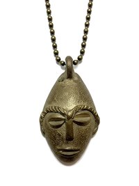 A brass pendant in the shape of a stylized human face with prominent brows, nose, and lips, hanging from a ball chain necklace on a white background.