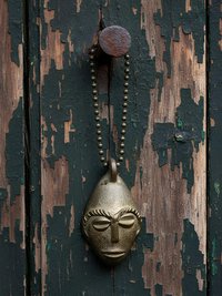 A bronze pendant shaped like a stylized human face hangs from a metal ball chain on a weathered, peeling wooden surface.