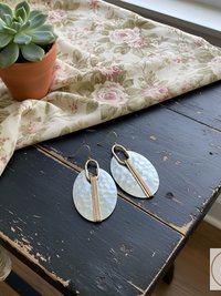 A pair of oval-shaped earrings with a hammered, pearlescent finish. The earrings have gold-colored accents and a vertical gold bar down the center. The earrings are displayed on a distressed black wooden surface.