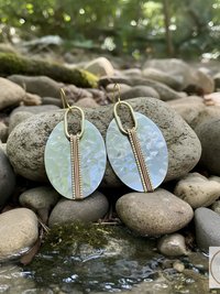 Oval-shaped earrings with a pearlescent finish, featuring a vertical copper and gold accent. The earrings have gold-colored ear wires and are displayed on a rock. The earrings are a fashion accessory.