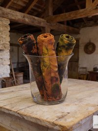 A clear glass vase holds three rolled tie dye fabric scarves. The scarves display a mix of colors, including brown, orange, and green, with a mottled pattern. The vase sits on a rustic, weathered wooden table.