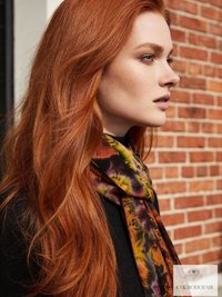 A woman with long red hair, wearing a colorful tie dye patterned scarf and a black top, stands in profile against a red brick wall.