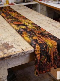 A long, rectangular scarf with a tie-dye pattern in shades of orange, yellow, and black. The scarf is draped across a distressed wooden table, showcasing the vibrant colors and unique design. The scarfs pattern creates a striking visual contrast.