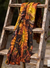 A tie-dye scarf in shades of orange, yellow, and black is draped over a rustic wooden ladder. The scarf's pattern features a blend of colors, creating a vibrant and artistic design. The ladder's aged appearance adds a vintage touch to the scene.