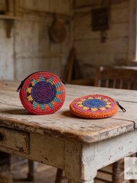 Two colorful, round, crocheted coin purses with zip closures sit on a rustic, worn wooden table in a cozy, vintage room with aged furniture and sunlight streaming in.